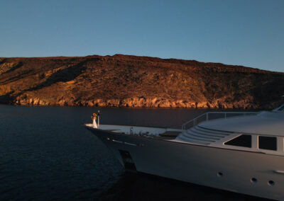 Drone footage of the exteriors of a yacht. A woman is standing in the bow during the golden hour.